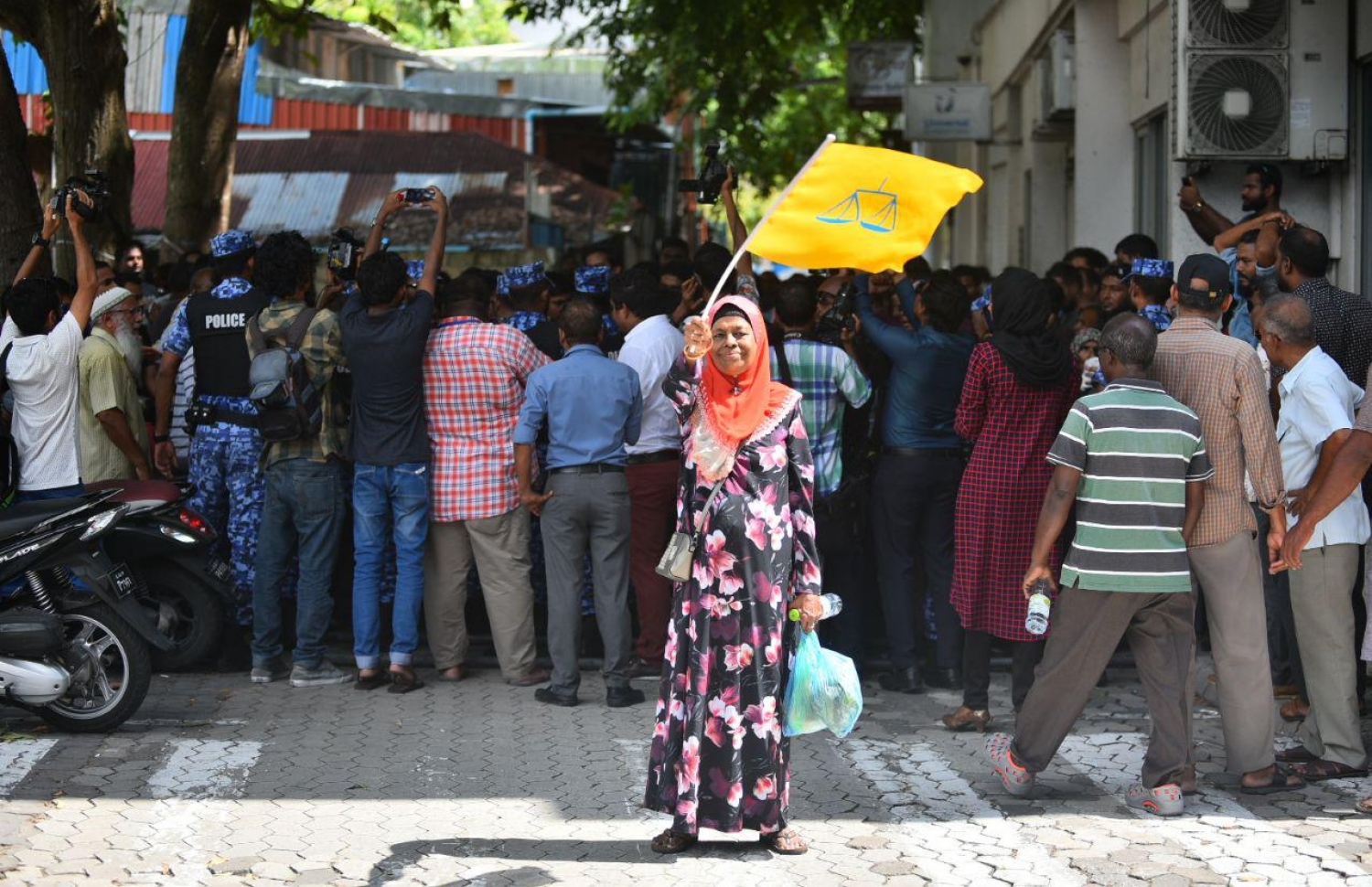 މާލެ: އޮކްޓޯބަރު 14، 2018: ސުޕްރީމް ކޯޓުގެ ހުޅަނގު ކަންމަތިން ކުރިމަތީގައި ހުންނަ ޔުނިވާސަލްގެ އިމާރާތުގެ ގޯޅިގަނޑަށް އެއްވެފައި ތިބި މީހުން ފަހަތަށް ޖެއްސުމަށް އެސްއޯ ފުލުހުންނާއި ސިފައިން ހަރަކާތްތެރިވާއިރު، މެދު އުމުރުގެ އަންހެނަކު އެމްޑީޕީ ދިދައެއް ހިފައިގެން.---ފޮޓޯ: އަހުމަދު ނިޝާތު/މިހާރު