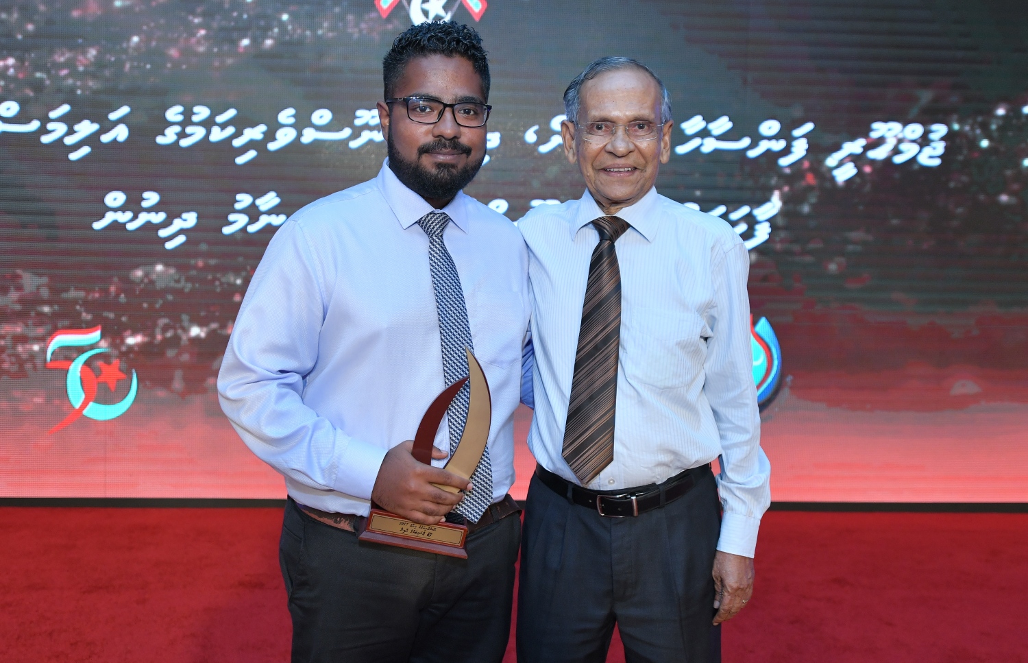 މާލެ: ޑިސެމްބަރު 11، 2018: ރާއްޖޭގެ ނޫސްވެރިކަމަށް 75 އަހަރު ފުރުމުގެ އަލިމަސްޔޫބީލް ފާހަގަ ކުރުމަށާއި ނޫސްވެރިކަމުގެ މިދިޔަ އަހަރުގެ އިނާމުތައް ދިނުމަށް މޯލްޑިވްސް މީޑިއާ ކައުންސިލާއި ހޯމް މިނިސްޓްރީ ގުޅިގެން ބޭއްވި ޖަލްސާގައި ނޫސްވެރިކަމުގެ ޝަރަފުގެ އަލިމަސް ގަލަން ހާސިލް ކުރެއްވި ޑރ. މުހައްމަދު ޒާހިރު ހުސައިން އާއެކު ފޮޓޯޖާނަލިސްޓުގެ އެވޯޑު ހާސިލްކުރި ރާއްޖެޓީވީގެ ޝަރުހާން ފޮޓޯއަކަށް ހުއްޓިގެން.---ފޮޓޯ: އަހުމަދު ނިޝާތު/މިހާރު