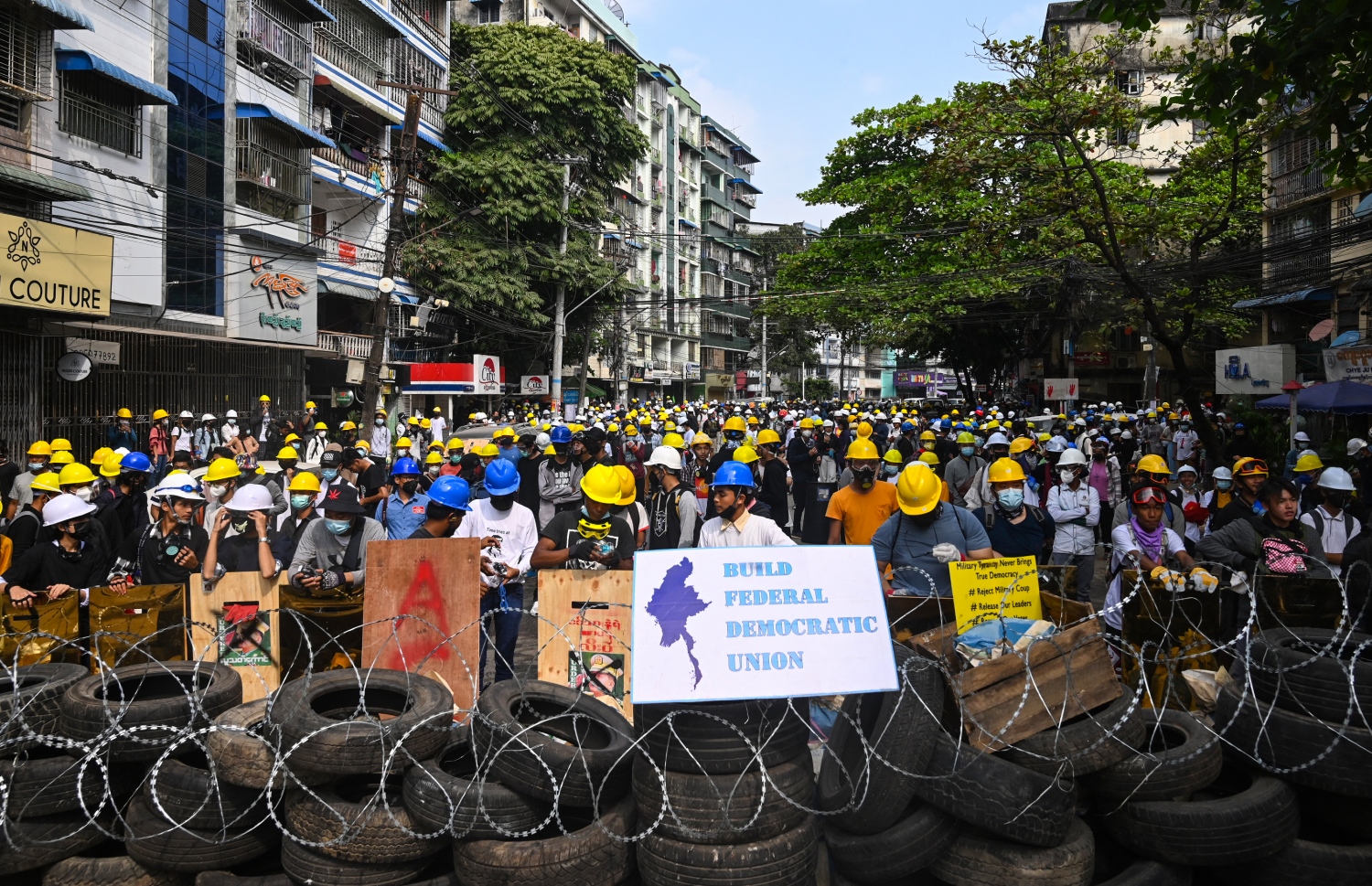 މިއަންމާގައި އިއްޔެ ކުރި މުޒާހަރާތަކުގައި ޔަންގޫންގައި ބެރިކޭޑްތަކުގެ ފަހަތުގައި ތިބިގެން އަޑުއުފުލަނީ. --ފޮޓޯ:އޭއެފްޕީ