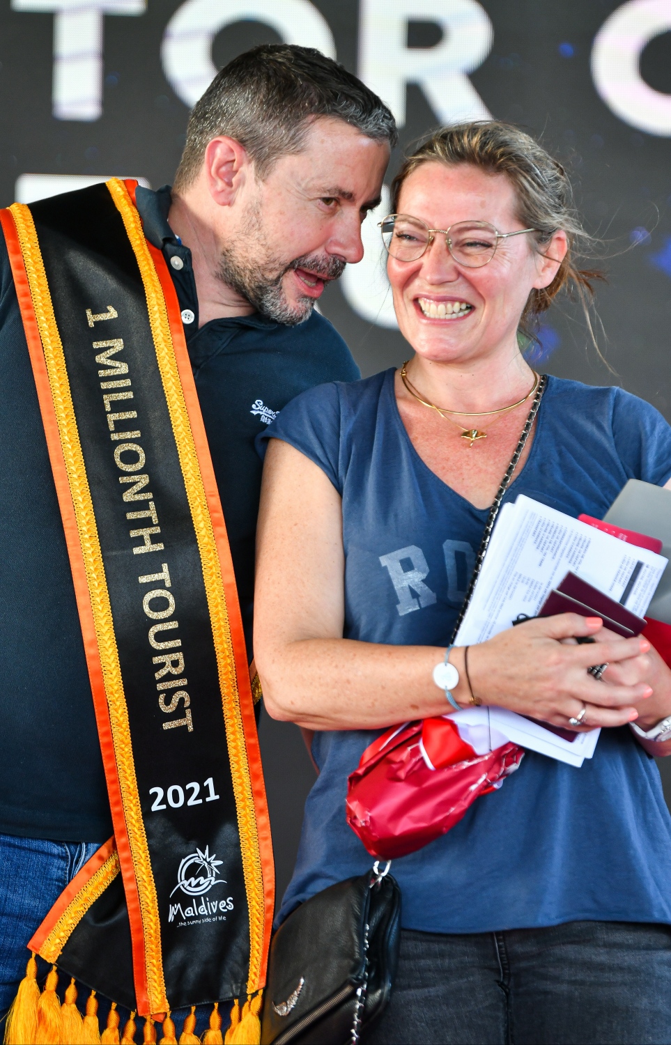 ވެލާނާ އެއާޕޯޓް: އޮކްޓޫބަރު 29، 2021- މި އަހަރު ރާއްޖެ އައި އެއް މިލިއަން ވަނަ ޓޫރިސްޓް ފްރާންސްގެ ވިންސެންޓް ވިކްޓާ ކޮރެން ޕިޓާއޫ އާއި އޭނާގެ އަންހެނުންގެ ޝަރަފުގައި ދިވެހި ސަގާފީ ހަރަކާތެއް ހުށަހަޅާއިރު، އެކަމާ އުފާކޮށް ދެ މީހުން ވާހަކަ ދައްކަނީ.--- ފޮޓޯ: ފަޔާޒު މޫސާ / މިހާރު