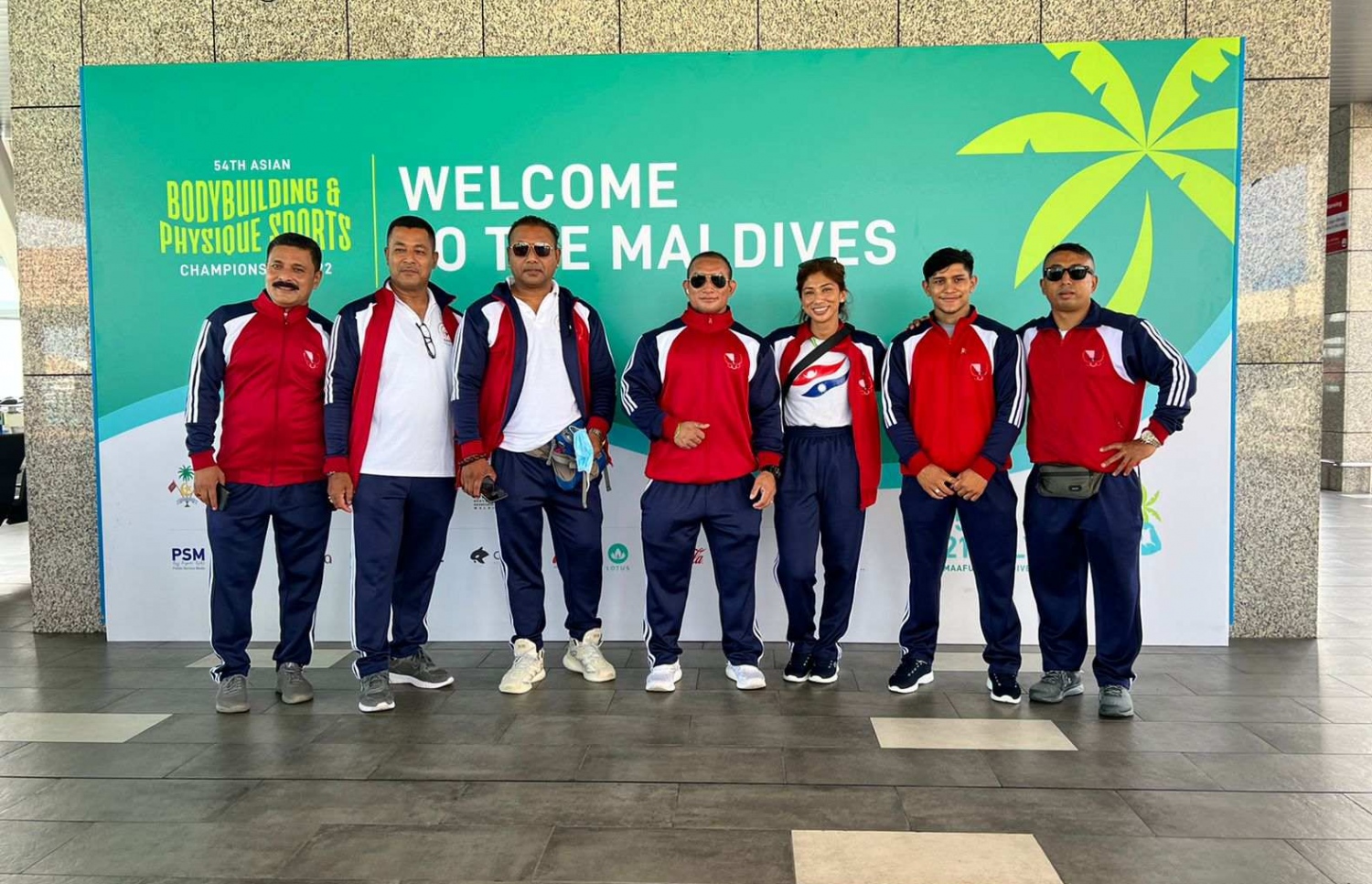ރާއްޖޭގައި ބާއްވާ 54 ވަނަ އޭޝިއަން ބޮޑީބިލްޑިންގް ޗެމްޕިއަންޝިޕްގައި ވާދަކުރަން އައި ނޭޕާލްގެ ޓީމުގެ ބައެއް އެތުލީޓުންނާއި އޮފިޝަލުން، މުބާރާތް ޕްރޮމޯޓް ކުރަން ވެލާނާ އިންޓަނޭޝަނަލް އެއާޕޯޓުގައި ހަރުކޮށްފައިވާ ބިލްބޯޑް ކައިރީގައި ފޮޓޯއަކަށް ހުއްޓިގެން.--- ފޮޓޯ: ބީބީއޭއެމް