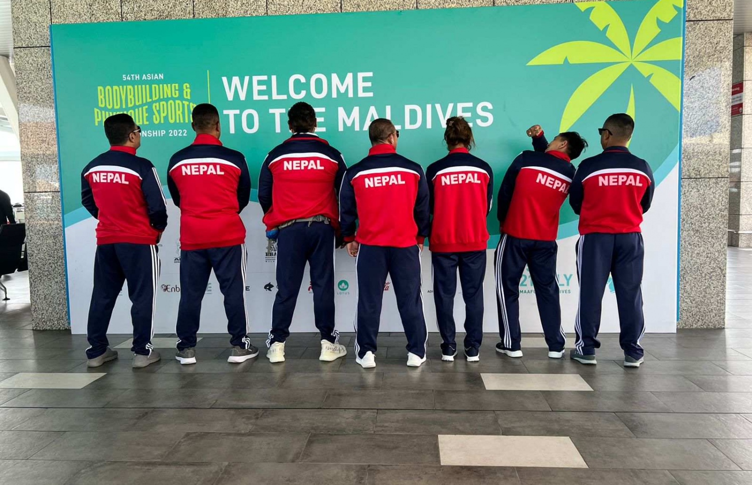 ރާއްޖޭގައި ބާއްވާ 54 ވަނަ އޭޝިއަން ބޮޑީބިލްޑިންގް ޗެމްޕިއަންޝިޕްގައި ވާދަކުރަން އައި ނޭޕާލްގެ ޓީމުގެ ބައެއް އެތުލީޓުންނާއި އޮފިޝަލުން، މުބާރާތް ޕްރޮމޯޓް ކުރަން ވެލާނާ އިންޓަނޭޝަނަލް އެއާޕޯޓުގައި ހަރުކޮށްފައިވާ ބިލްބޯޑް ކައިރީގައި ފޮޓޯއަކަށް ހުއްޓިގެން.--- ފޮޓޯ: ބީބީއޭއެމް