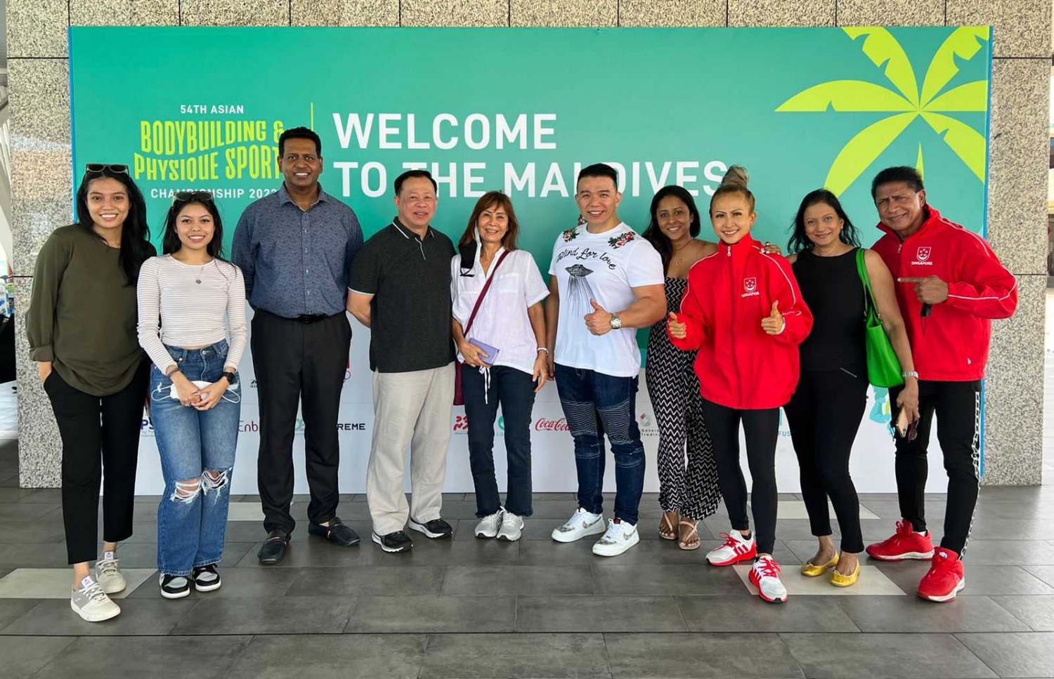 ރާއްޖޭގައި ބާއްވާ 54 ވަނަ އޭޝިއަން ބޮޑީބިލްޑިންގް ޗެމްޕިއަންޝިޕްގައި ވާދަކުރަން އައި ސިންގަޕޫރުގެ ޓީމުގެ ބައެއް އެތުލީޓުން، މުބާރާތް ޕްރޮމޯޓް ކުރަން ވެލާނާ އިންޓަނޭޝަނަލް އެއާޕޯޓުގައި ހަރުކޮށްފައިވާ ބިލްބޯޑް ކައިރީ ފޮޓޯއަކަށް ހުއްޓިގެން.--- ފޮޓޯ: ބީބީއޭއެމް