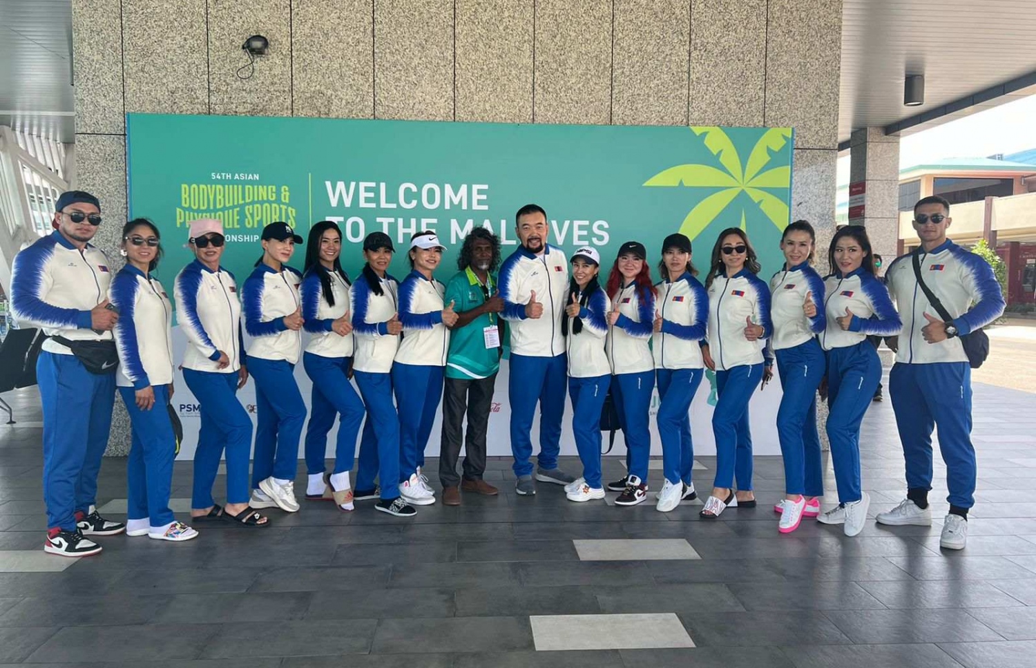 ރާއްޖޭގައި ބާއްވާ 54 ވަނަ އޭޝިއަން ބޮޑީބިލްޑިންގް ޗެމްޕިއަންޝިޕްގައި ވާދަކުރަން އައި މޮންގޯލިއާގެ ޓީމުގެ އެތުލީތުންނާއި އޮފިޝަލުން، މުބާރާތް ޕްރޮމޯޓް ކުރަން ވެލާނާ އިންޓަނޭޝަނަލް އެއާޕޯޓުގައި ހަރުކޮށްފައިވާ ބިލްބޯޑް ކައިރީގައި ފޮޓޯއަކަށް ހުއްޓިގެން.--- ފޮޓޯ: ބީބީއޭއެމް