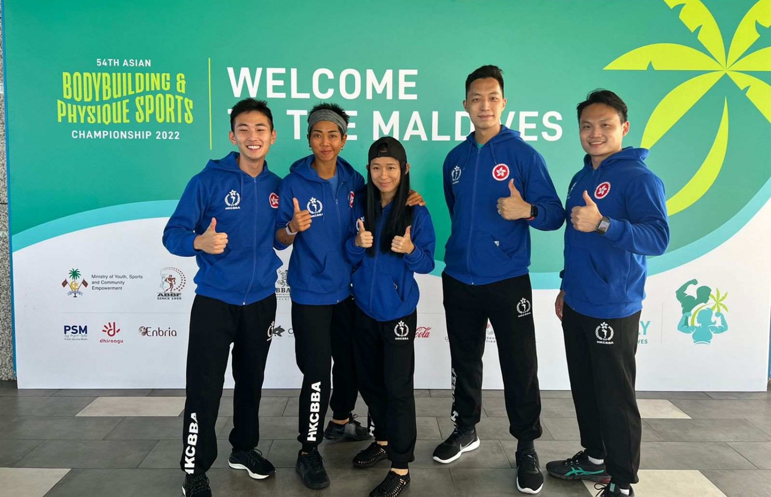 ރާއްޖޭގައި ބާއްވާ 54 ވަނަ އޭޝިއަން ބޮޑީބިލްޑިންގް ޗެމްޕިއަންޝިޕްގައި ވާދަކުރަން އައި ހޮންކޮންގްގެ ޓީމުގެ ބައެއް އެތުލީޓުން، މުބާރާތް ޕްރޮމޯޓް ކުރަން ވެލާނާ އިންޓަނޭޝަނަލް އެއާޕޯޓުގައި ހަރުކޮށްފައިވާ ބިލްބޯޑު ކައިރީގައި ފޮޓޯއަކަށް ހުއްޓިގެން.---ފޮޓޯ: ބީބީއޭއެމް