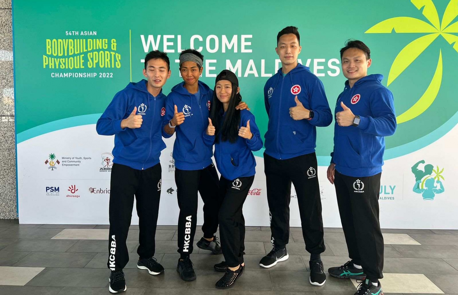 ރާއްޖޭގައި ބާއްވާ 54 ވަނަ އޭޝިއަން ބޮޑީބިލްޑިންގް ޗެމްޕިއަންޝިޕްގައި ވާދަކުރަން އައި ހޮންކޮންގްގެ ޓީމުގެ ބައެއް އެތުލީޓުންނާއި އޮފިޝަލުން، މުބާރާތް ޕްރޮމޯޓް ކުރަން ވެލާނާ އިންޓަނޭޝަނަލް އެއާޕޯޓުގައި ހަރުކޮށްފައިވާ ބިލްބޯޑް ކައިރީގައި ފޮޓޯއަކަށް ހުއްޓިގެން.--- ފޮޓޯ: ބީބީއޭއެމް