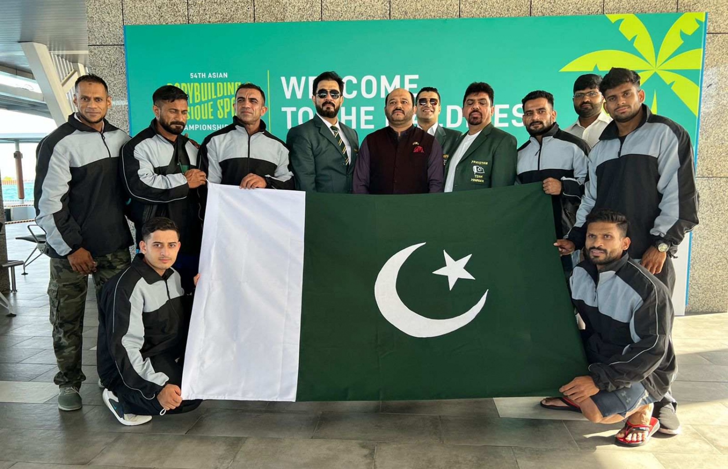 ރާއްޖޭގައި ބާއްވާ 54 ވަނަ އޭޝިއަން ބޮޑީބިލްޑިންގް ޗެމްޕިއަންޝިޕްގައި ވާދަކުރަން އައި ޕާކިސްތާނުގެ ޓީމުގެ ބައެއް އެތުލީޓުންނާއި އޮފިޝަލުން، މުބާރާތް ޕްރޮމޯޓް ކުރަން ވެލާނާ އިންޓަނޭޝަނަލް އެއާޕޯޓުގައި ހަރުކޮށްފައިވާ ބިލްބޯޑް ކައިރީގައި ފޮޓޯއަކަށް ހުއްޓިގެން.--- ފޮޓޯ: ބީބީއޭއެމް