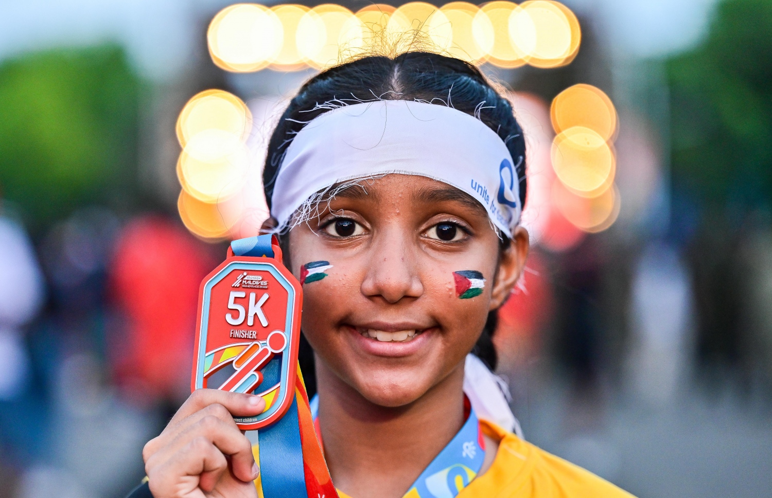 ރާއްޖޭގައި ބާއްވާ އެންމެ ބޮޑު ދުވުމުގެ އިވެންޓް ކަމަށްވާ ދިރާގު މޯލްޑިވްސް ރޯޑް ރޭހުގެ ބައިވެރިވި ކުޑަ ކުއްޖަކު މެޑެލްއާއެކު ފޮޓޯއަކަށް ހުއްޓިލައިގެން ހުރިއިރު މޫނުގައި ފަލަސްތީންގެ ދިދަ ކުރަހާފަ -- ފޮޓޯ: ފަޔާޒު މޫސާ | މިހާރު