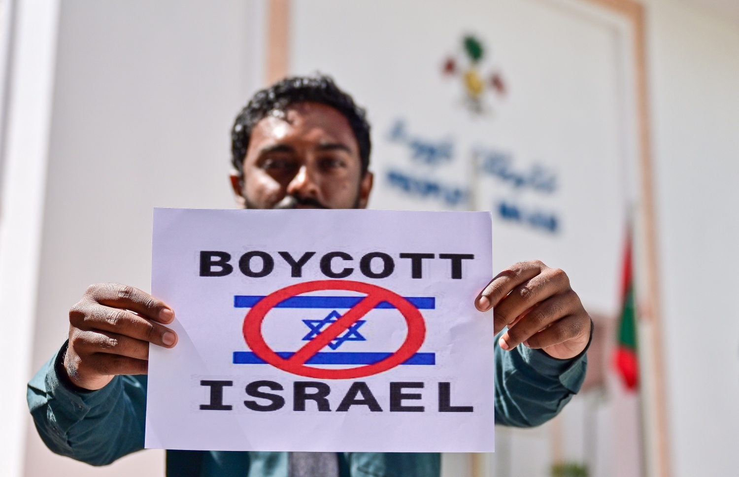 ރައްޔިތުންގެ މަޖިލީހުގެ އިދާރާ ކުރިމަތީ ބޭއްވި އިހުތިޖާޖުގައި "ބޮއިކޮޓް އިޒްރާއިލް" ލިޔެފައިވާ ޕޯސްޓަރެއް ހިފައިގެން މަޖިލިސް ދޮރުމަތީގައި މީހަކު މަޑުކޮށްލައިގެން.--- ފޮޓޯ: ފަޔާޒު މޫސާ | މިހާރު