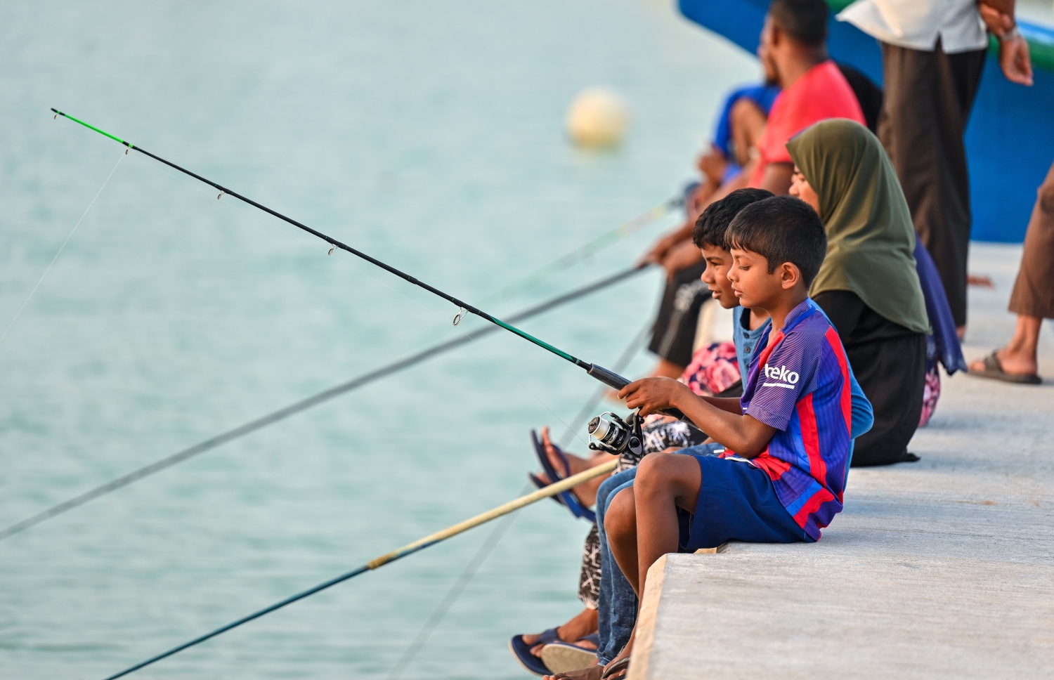 ކުޑަކުދިންތަކެއް އެ ކުދިންގެ ބެލެނިވެރިންނާ އެކު ރަށުގެ ބަނދަރު ސަރަހައްދުގައި މުށިމަސް ބާނަނީ: މުށި މަސް ބާނައިގެން ކައްކައި ހިއްކައި ރިހާކުރު ހަދާ މީހުން ވެސް މި ރަށުގައި އުޅޭ. -- ފޮޓޯ: ފަޔާޒު މޫސާ | މިހާރު