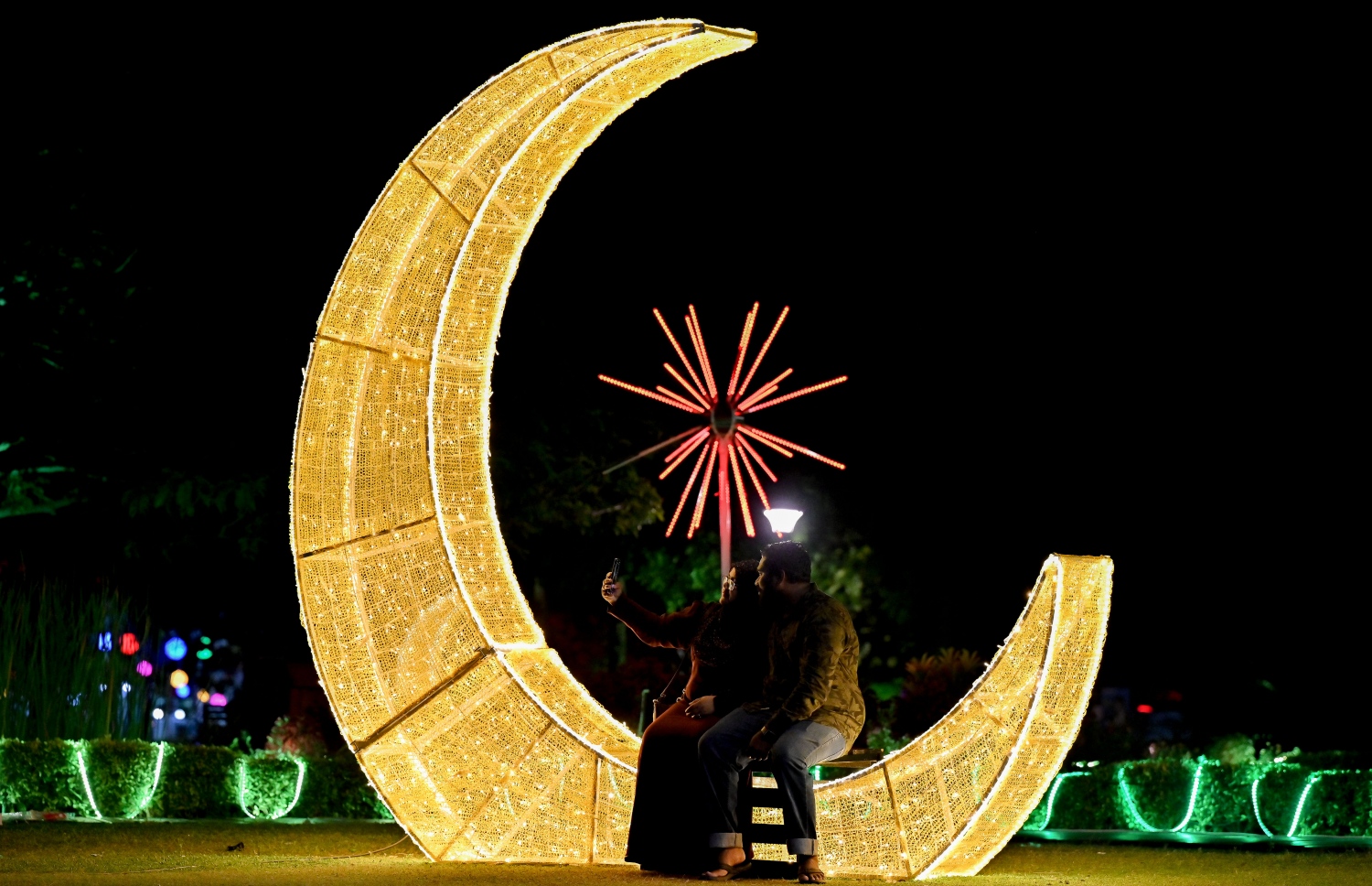 ފިތުރު އީދަށް ހުޅުމާލޭގެ އެކި ސަރަހައްދުތައް ކުލަ ބޮކިތަކާއި ޑިޒައިންތަކުން ޒީނަތްތެރިކޮށްފައި ވާއިރު ހުޅުމާލެ ސެންޓްރަލް ޕާކްގައި ދިއްލާފައިވާ ހަނދުފަޅިއެއް ގޮތަށް ހުރި ތަނެއްގައި އިށީނދެ ތިބެގެން ދެ މީހަކު ފޮޓޯ ނަގަނީ.-- ފޮޓޯ: ނިޝާން އަލީ | މިހާރު